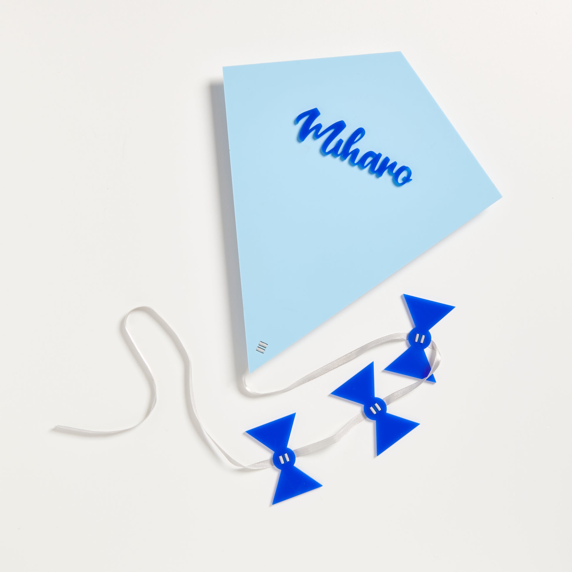 A photo of a kite made from 3mm pastel blue acrylic and topped with the name Miharo  in Blue gloss acrylic. The kite also has a silver satin ribbon and 3 blue acrylic bows attached..