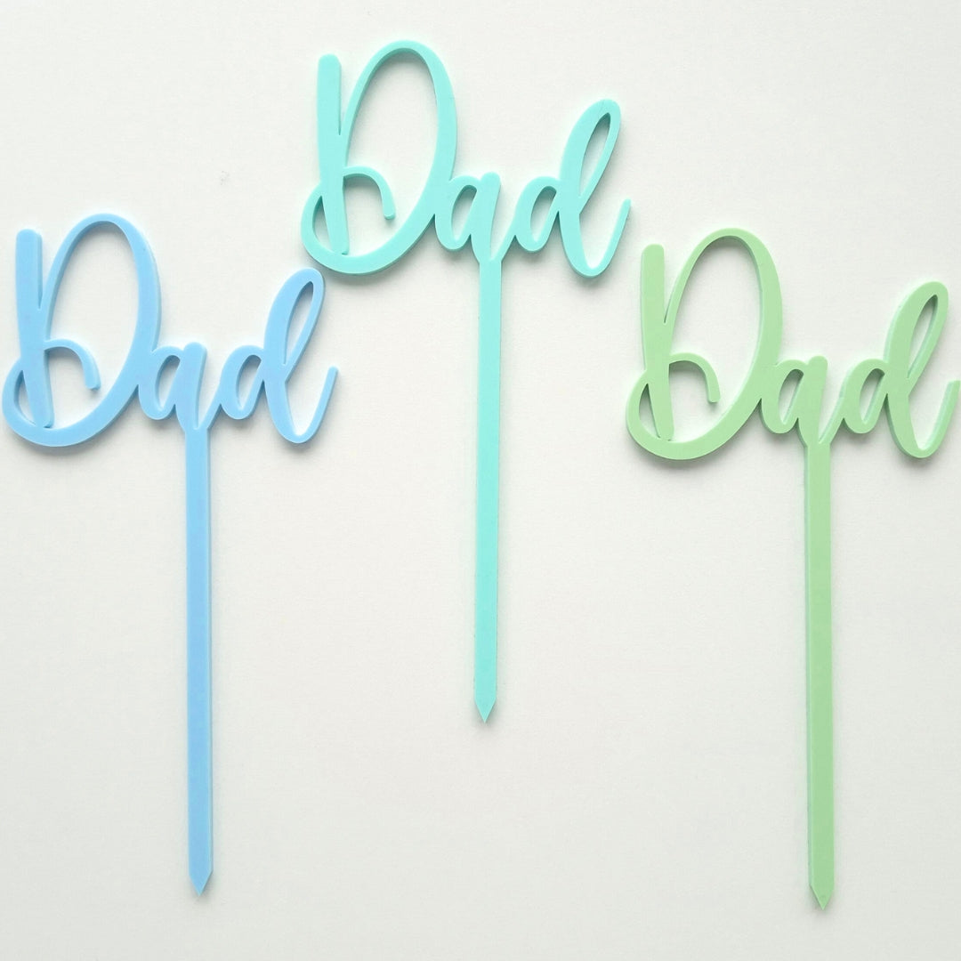 Three 'Dad' cake toppers in blue, green, and light blue on a white background