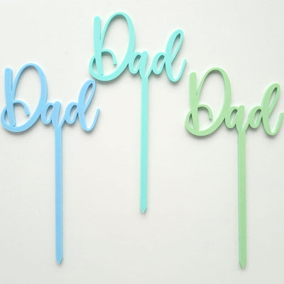Three 'Dad' cake toppers in blue, green, and light blue on a white background