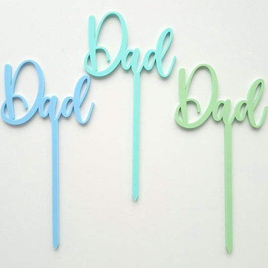 Three 'Dad' cake toppers in blue, green, and light blue on a white background